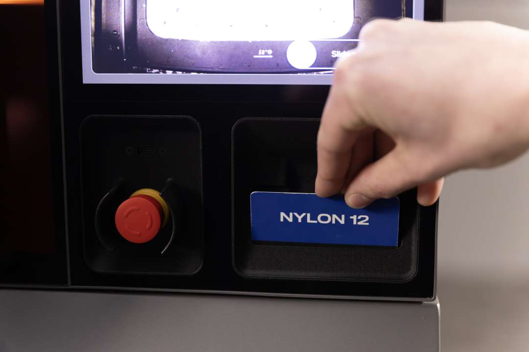 Рука вставляє синю NFC-картку матеріалу Nylon 12 у зчитувач Formlabs Fuse X1; зліва видно червоний аварійний стоп