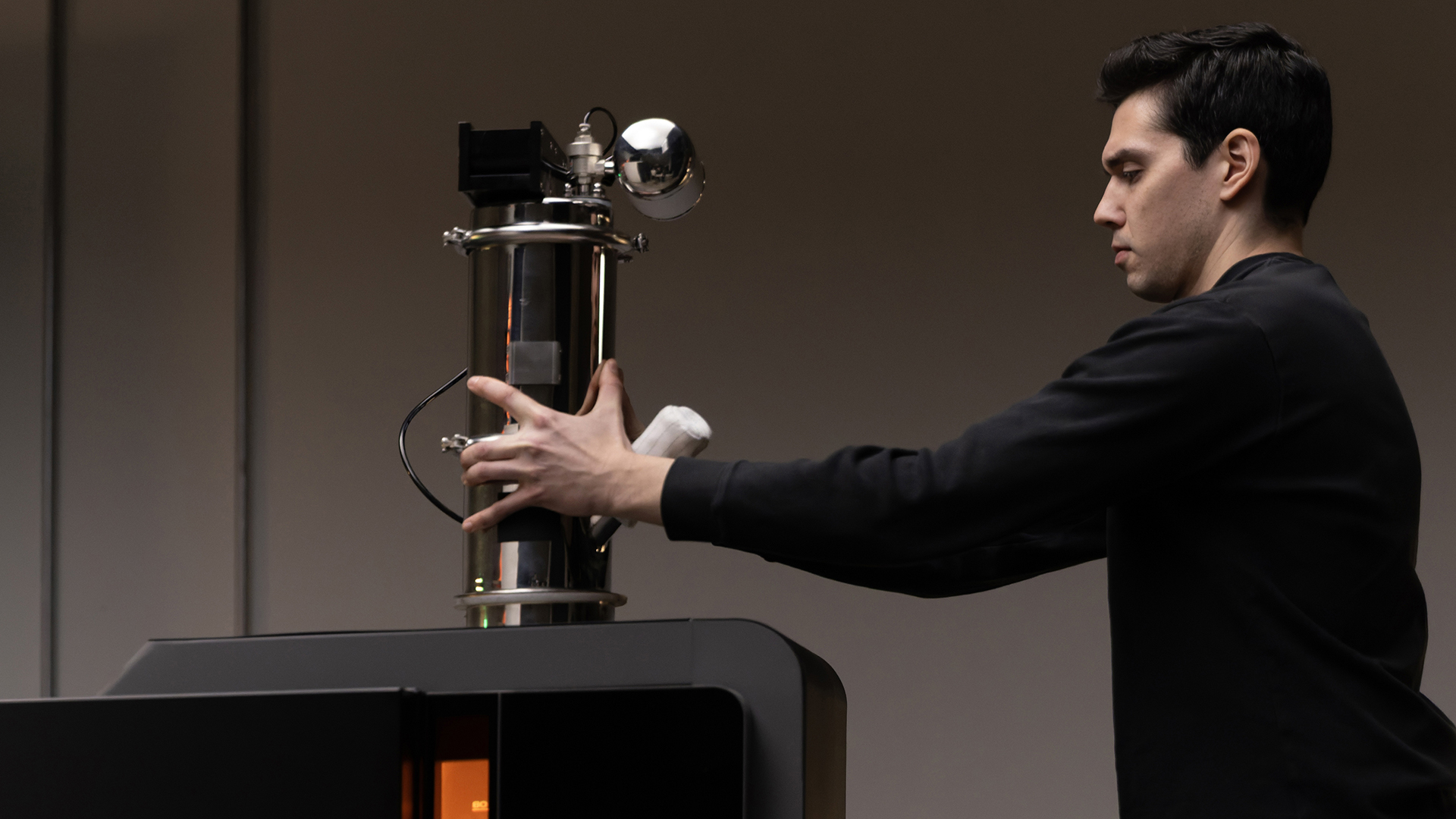 Фахівець у чорному светрі встановлює Formlabs Fuse X1 Vacuum Conveyor на верхню частину SLS-принтера Fuse X1, крізь переднє вікно якого видно оранжеве підсвічування камери